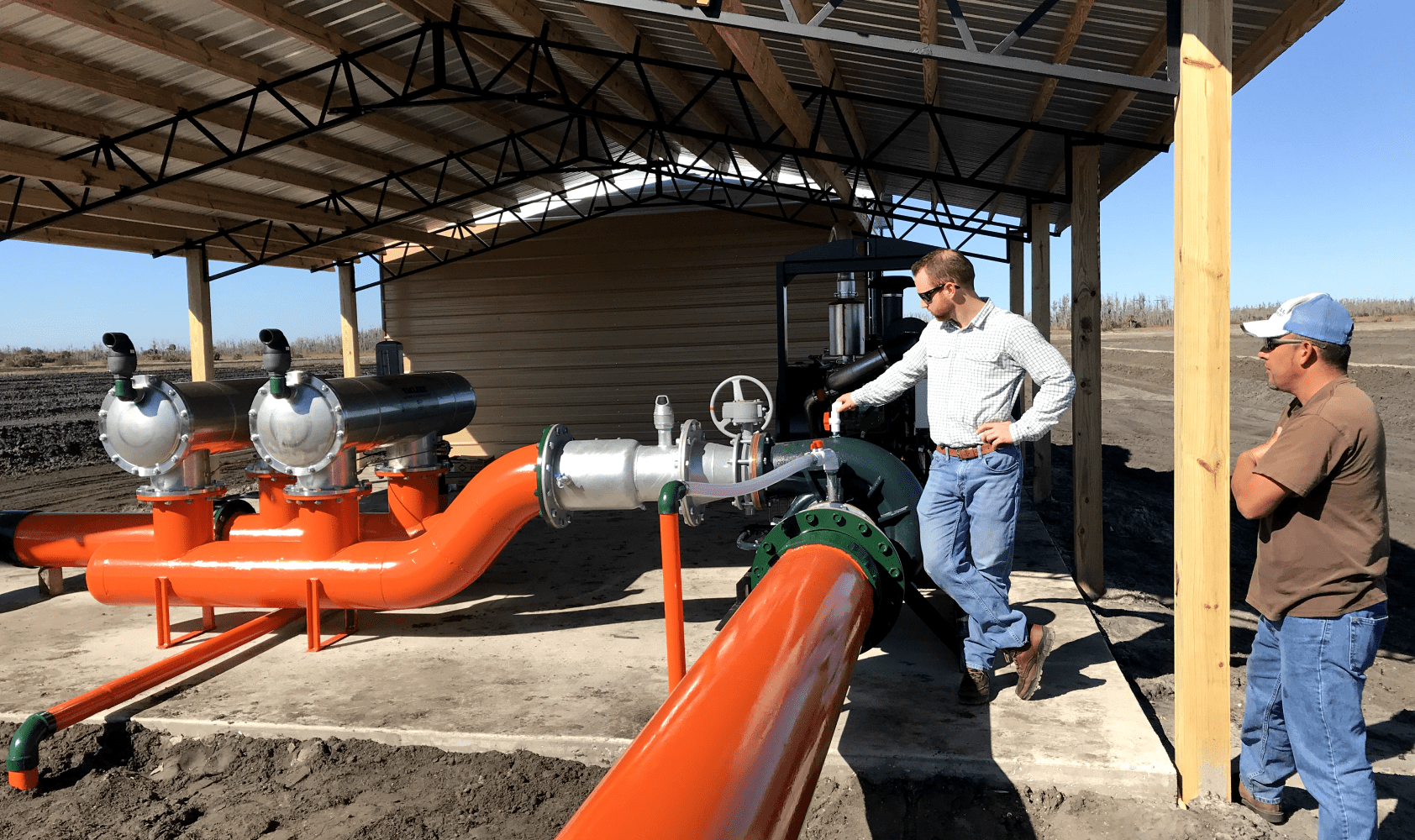 Two men standing next to large orange pipes under a metal roof.